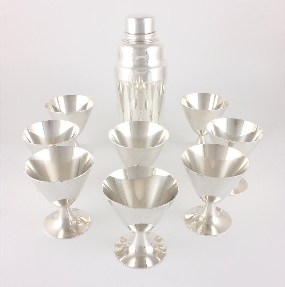 A silver-plated Art Deco cocktail shaker accompanied by eight matching conical goblet glasses arranged on a white background.