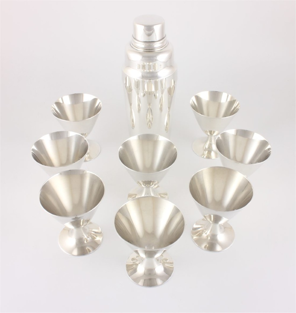 A silver-plated Art Deco cocktail shaker accompanied by eight matching conical goblet glasses arranged on a white background. View III