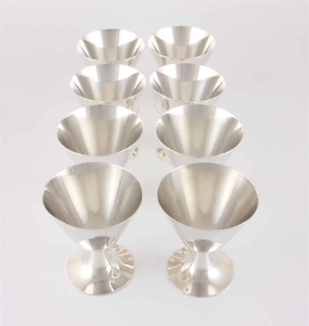 A silver-plated Art Deco cocktail shaker accompanied by eight matching conical goblet glasses arranged on a white background. View V