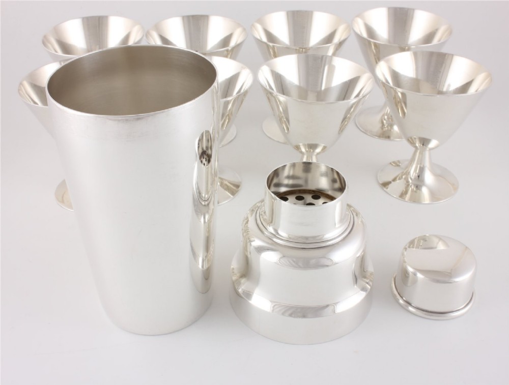 A silver-plated Art Deco cocktail shaker accompanied by eight matching conical goblet glasses arranged on a white background. View VI