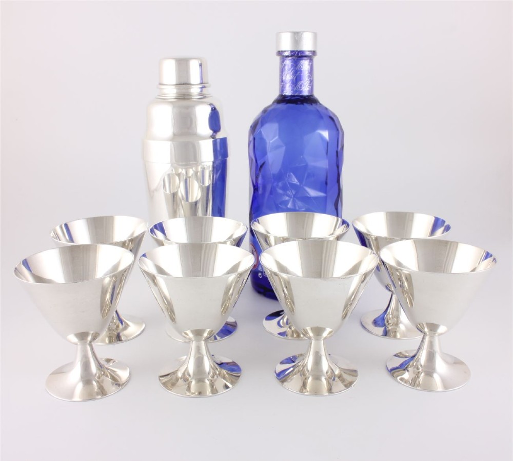 A silver-plated Art Deco cocktail shaker accompanied by eight matching conical goblet glasses arranged on a white background. View XI