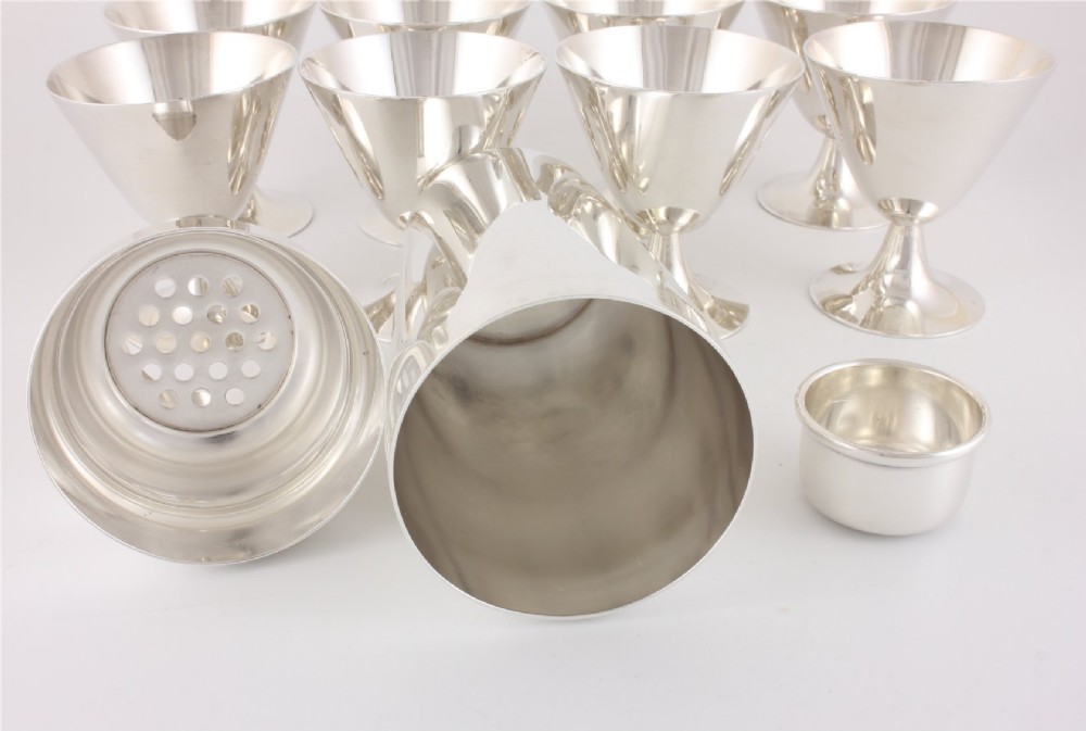 A silver-plated Art Deco cocktail shaker accompanied by eight matching conical goblet glasses arranged on a white background. View XII