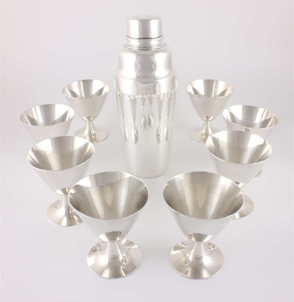 A silver-plated Art Deco cocktail shaker accompanied by eight matching conical goblet glasses arranged on a white background. View XIII