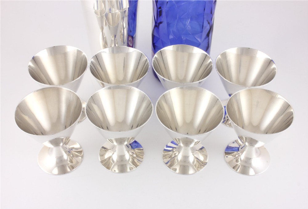 A silver-plated Art Deco cocktail shaker accompanied by eight matching conical goblet glasses arranged on a white background. View XIV