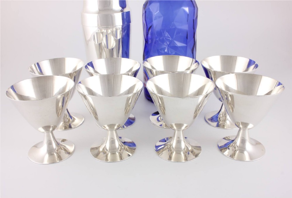 A silver-plated Art Deco cocktail shaker accompanied by eight matching conical goblet glasses arranged on a white background. View XV