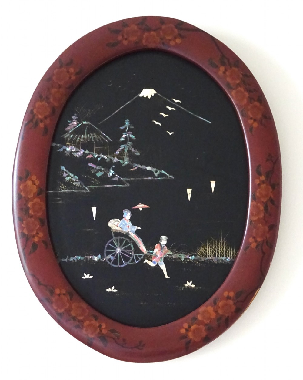 An oval red-lacquered framed artwork depicting a Japanese scene with Mount Fuji and a rickshaw in mother-of-pearl and abalone shell inlay against a black background.