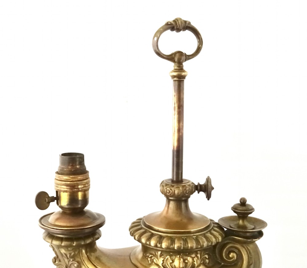 An ornate late Victorian brass oil lamp featuring a weighted circular base, central stem, detailed reservoir with relief carving, and a top carrying ring handle. View VIII