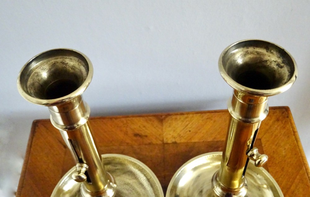 A pair of late Victorian polished brass ejector candlesticks featuring turned stems, side slider knobs, and wide dished bases. View V