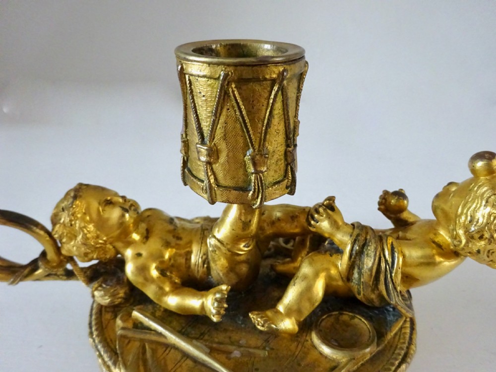 A gilt bronze candleholder featuring two reclining cherub figures holding a drum-shaped socket, with a french horn handle and tasselled circular base. View III