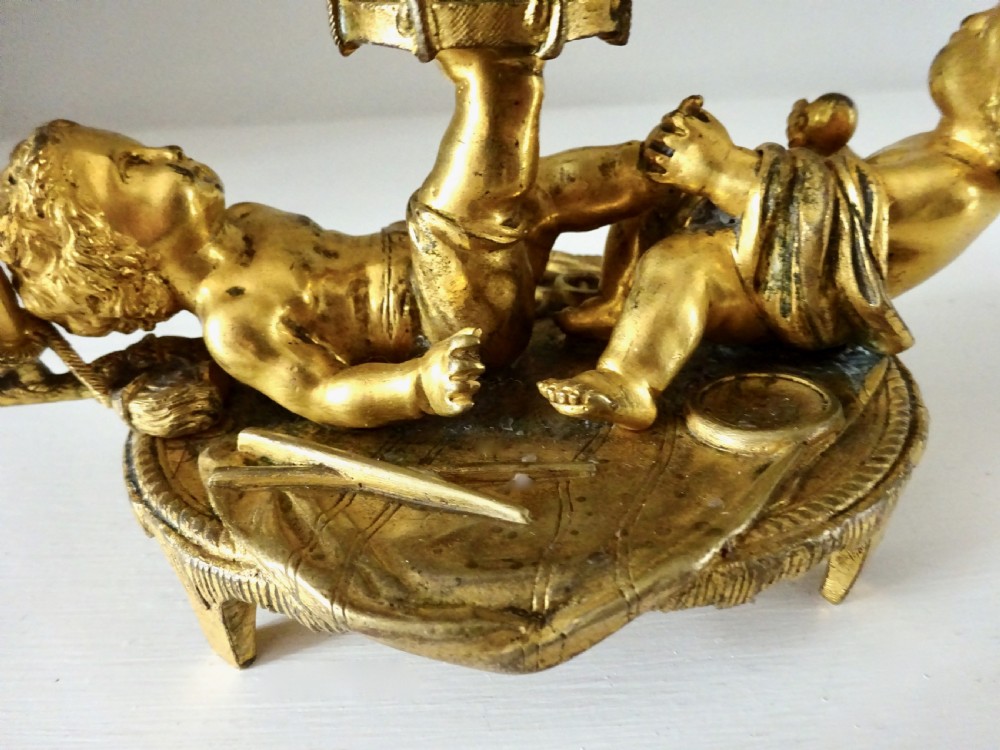 A gilt bronze candleholder featuring two reclining cherub figures holding a drum-shaped socket, with a french horn handle and tasselled circular base. View IV
