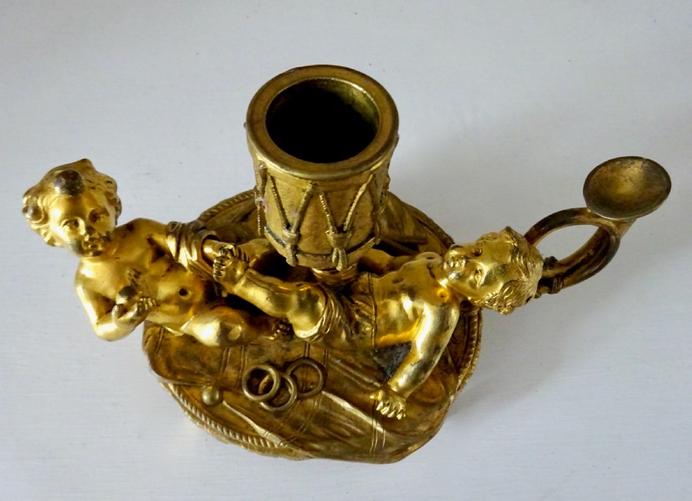 A gilt bronze candleholder featuring two reclining cherub figures holding a drum-shaped socket, with a french horn handle and tasselled circular base. View VI