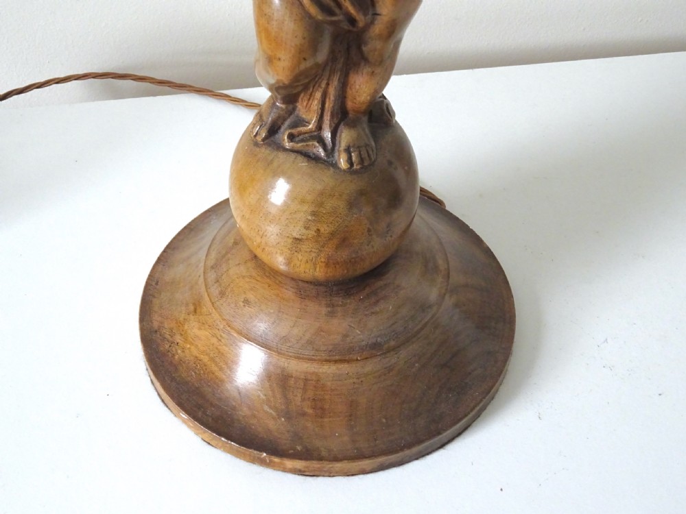 A hand-carved oak table lamp featuring a cherub figure standing on an orb base and holding a fluted stem, topped with a neutral fabric shade. View VI