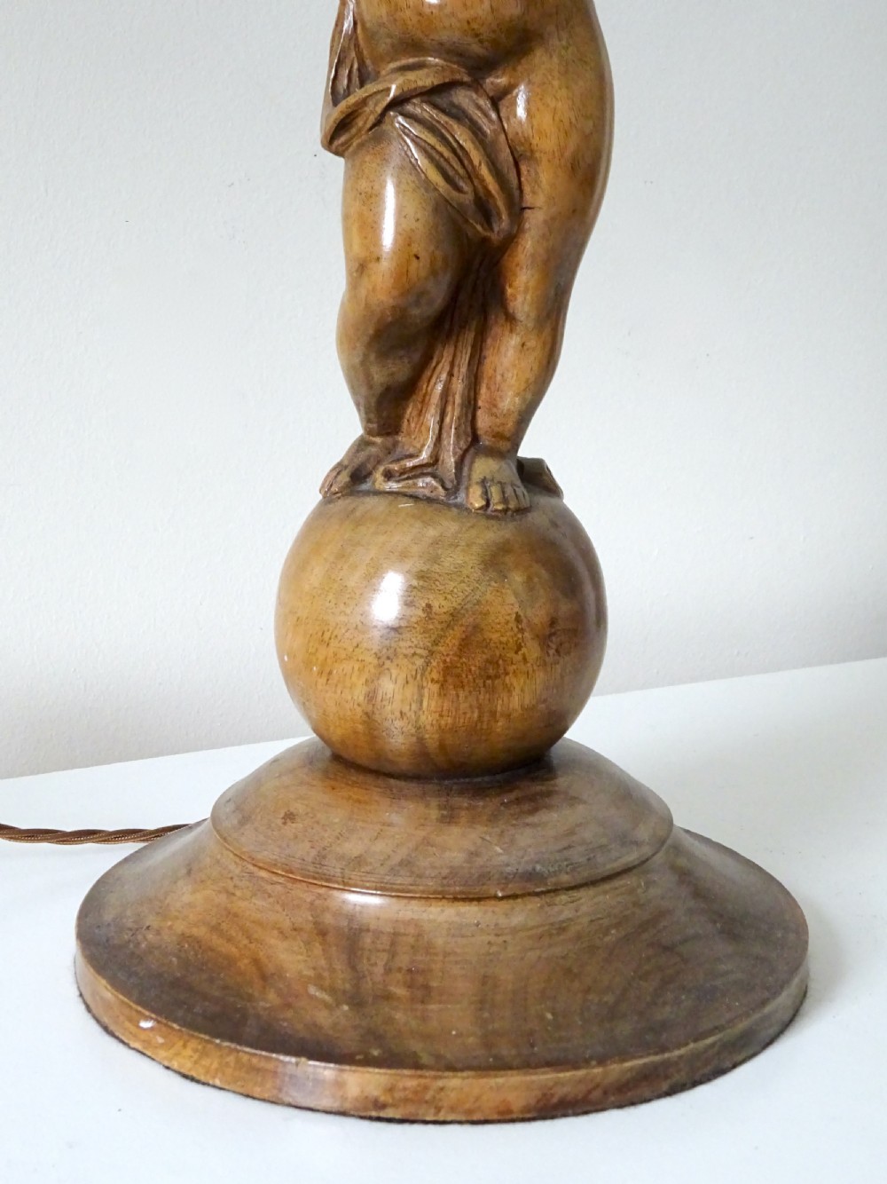A hand-carved oak table lamp featuring a cherub figure standing on an orb base and holding a fluted stem, topped with a neutral fabric shade. View IX