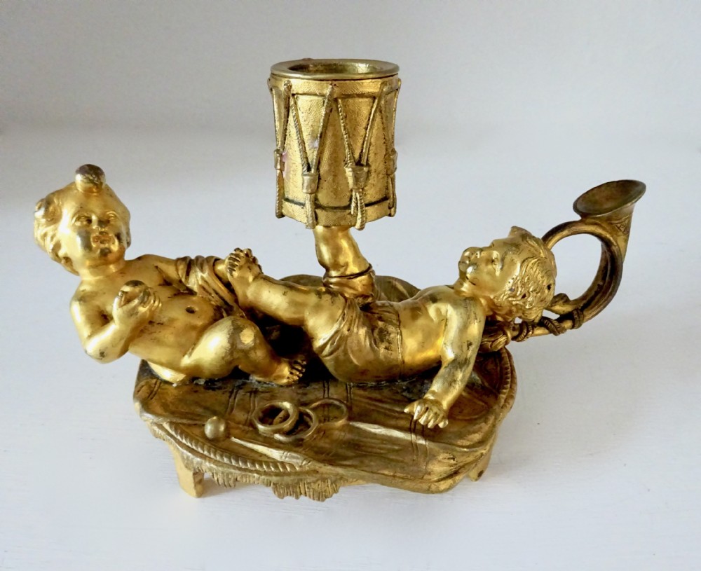 A gilt bronze candleholder featuring two reclining cherub figures holding a drum-shaped socket, with a french horn handle and tasselled circular base. View V