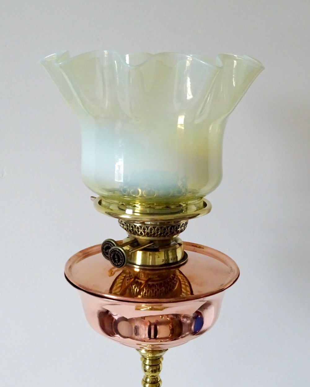 An Arts and Crafts oil lamp featuring a square brass base, a slender turned column stem, a polished copper reservoir, and a pale yellow frilled vaseline glass shade. View III
