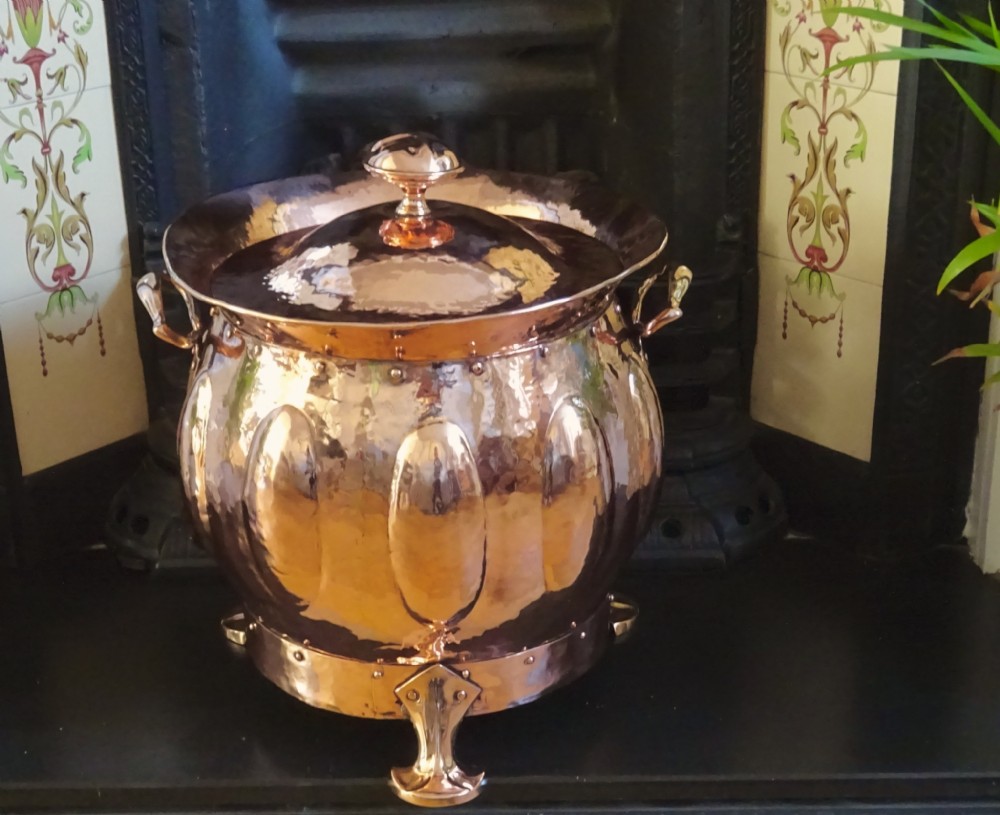 A polished hand-hammered copper log bin featuring a fitted lid with a central finial, repousse lobed sides, side handles, and decorative tripod feet. View IV
