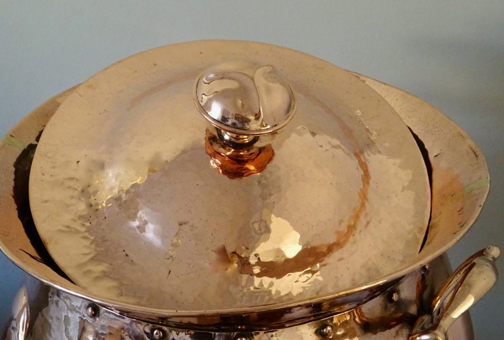 A polished hand-hammered copper log bin featuring a fitted lid with a central finial, repousse lobed sides, side handles, and decorative tripod feet. View VI