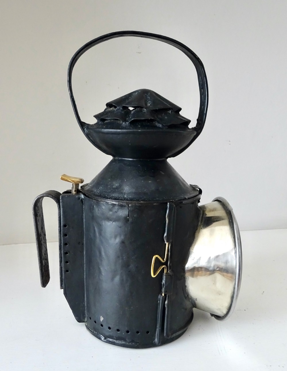 A black painted metal railway oil lantern featuring a hinged glass lens door, carrying handle, and internal brass oil burner. View VII
