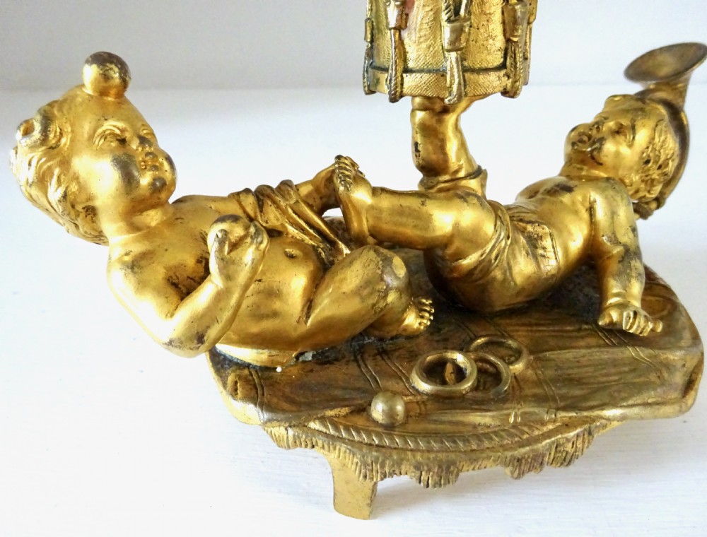 A gilt bronze candleholder featuring two reclining cherub figures holding a drum-shaped socket, with a french horn handle and tasselled circular base. View II