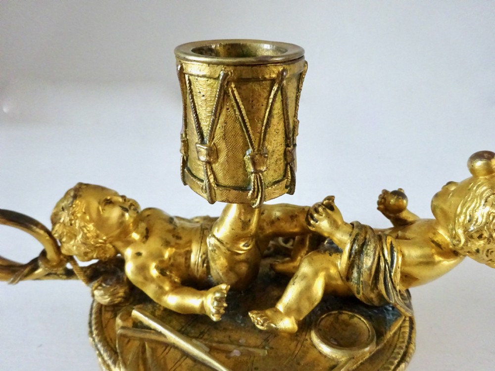 A gilt bronze candleholder featuring two reclining cherub figures holding a drum-shaped socket, with a french horn handle and tasselled circular base. View IX