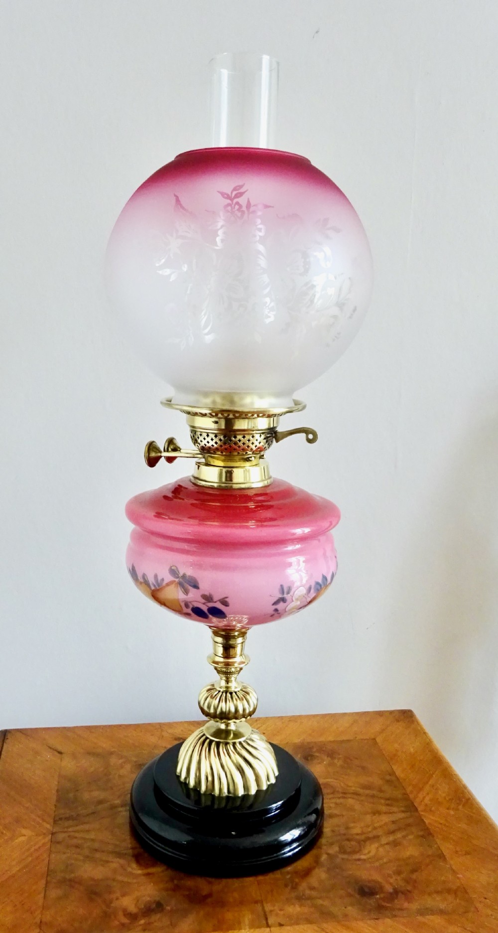 An early twentieth-century brass oil lamp featuring a pink hand-painted glass reservoir, an etched floral glass globe shade, a clear glass chimney, and a black stepped base.