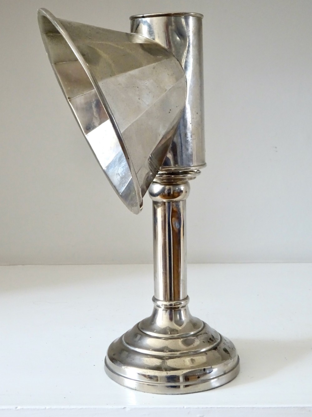 A nickel-plated antique candle lamp featuring a stepped circular base, tubular stem, and a curved fan-like reflector hood attached near the candle nozzle. View III