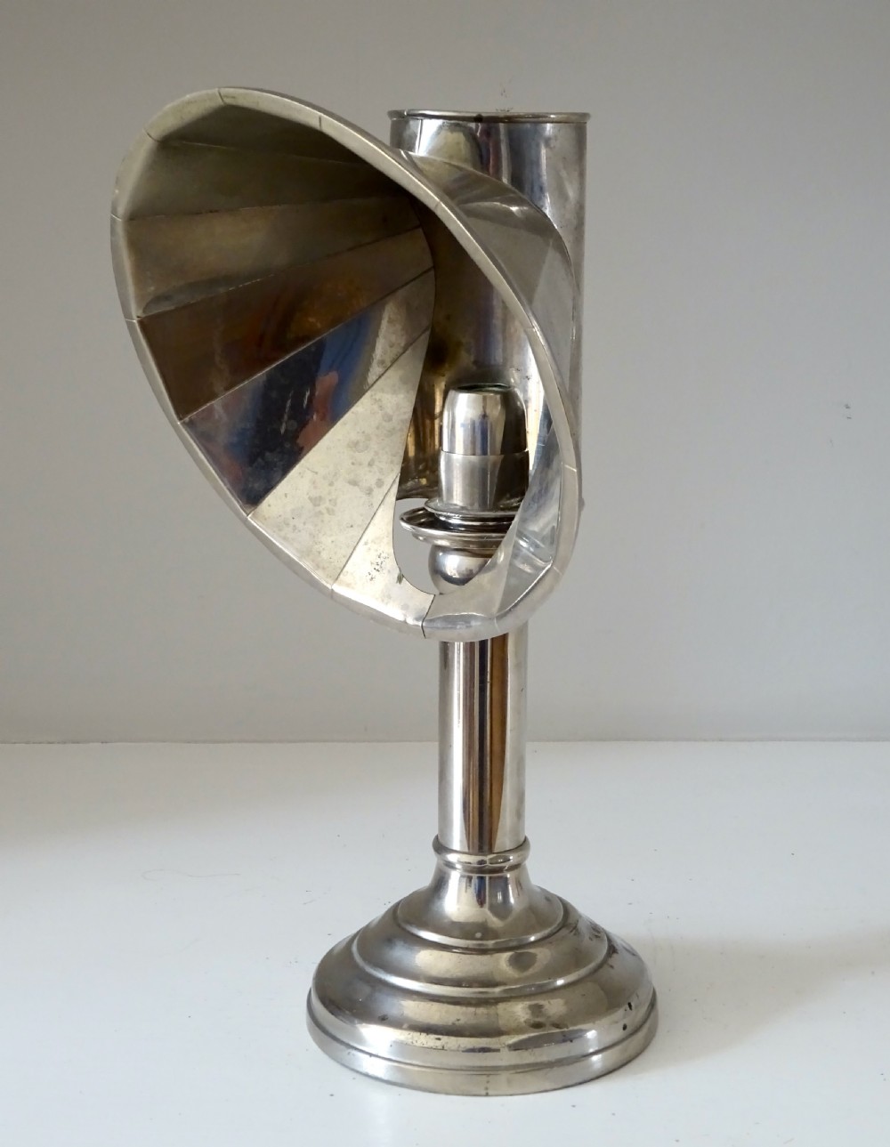 A nickel-plated antique candle lamp featuring a stepped circular base, tubular stem, and a curved fan-like reflector hood attached near the candle nozzle.