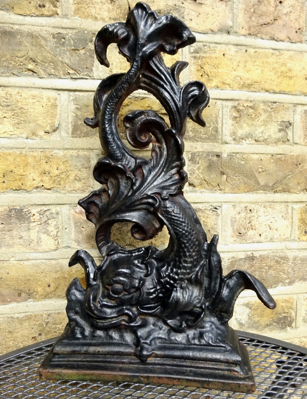 A heavy black cast iron doorstop featuring a mythological dolphin intertwined with acanthus scrollwork set upon a rectangular molded base.