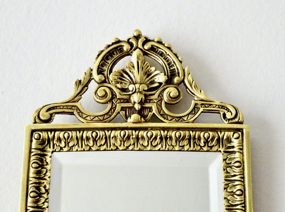 An ornate rectangular cast bronze wall mirror featuring a bevelled glass plate, classical scroll cresting, embossed repeating frame pattern, and a mask apron at the bottom. View II
