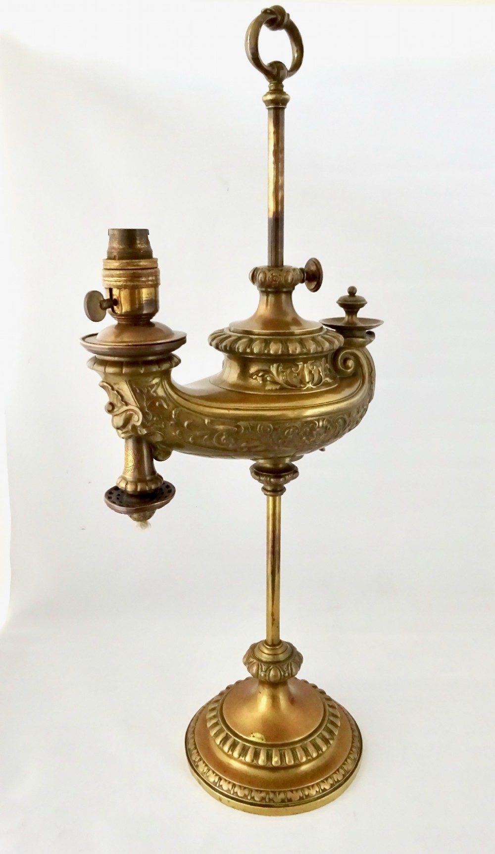 An ornate late Victorian brass oil lamp featuring a weighted circular base, central stem, detailed reservoir with relief carving, and a top carrying ring handle. View V