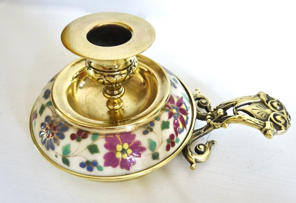 An Edwardian brass chamberstick with an ornate cast handle and a circular base decorated with floral enamel patterns. View IV