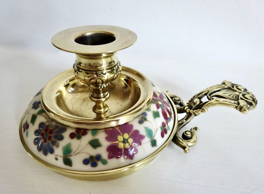 An Edwardian brass chamberstick with an ornate cast handle and a circular base decorated with floral enamel patterns.