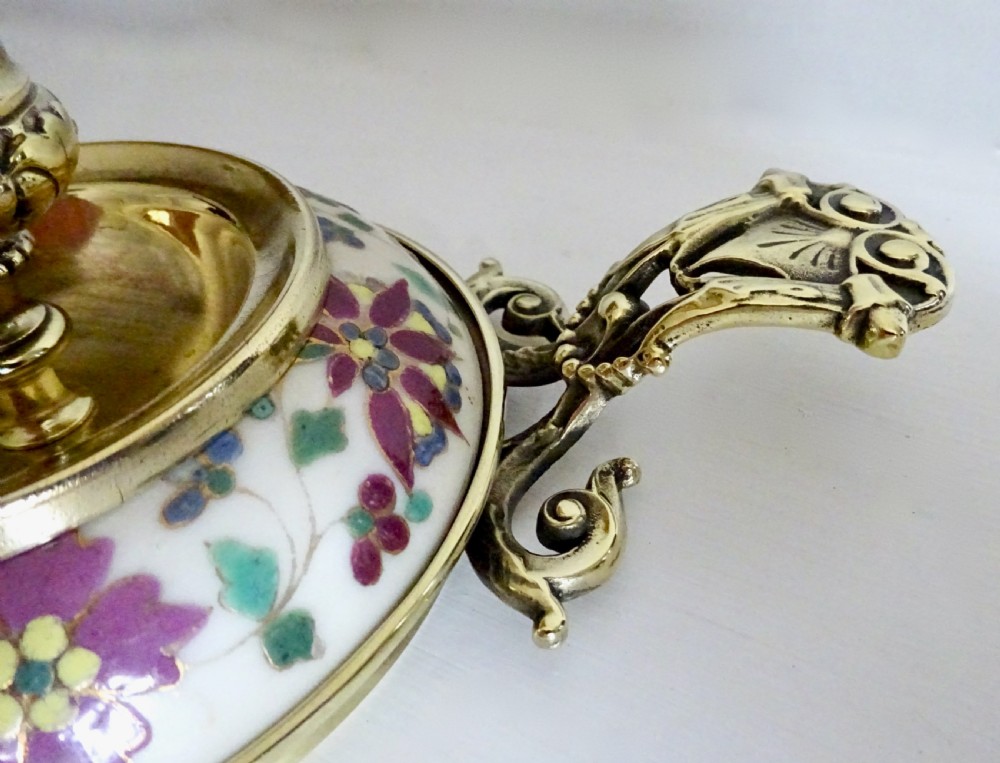 An Edwardian brass chamberstick with an ornate cast handle and a circular base decorated with floral enamel patterns. View III