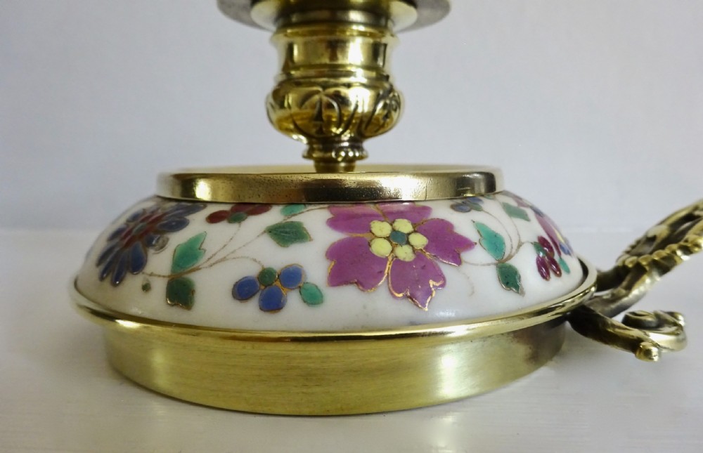 An Edwardian brass chamberstick with an ornate cast handle and a circular base decorated with floral enamel patterns. View V