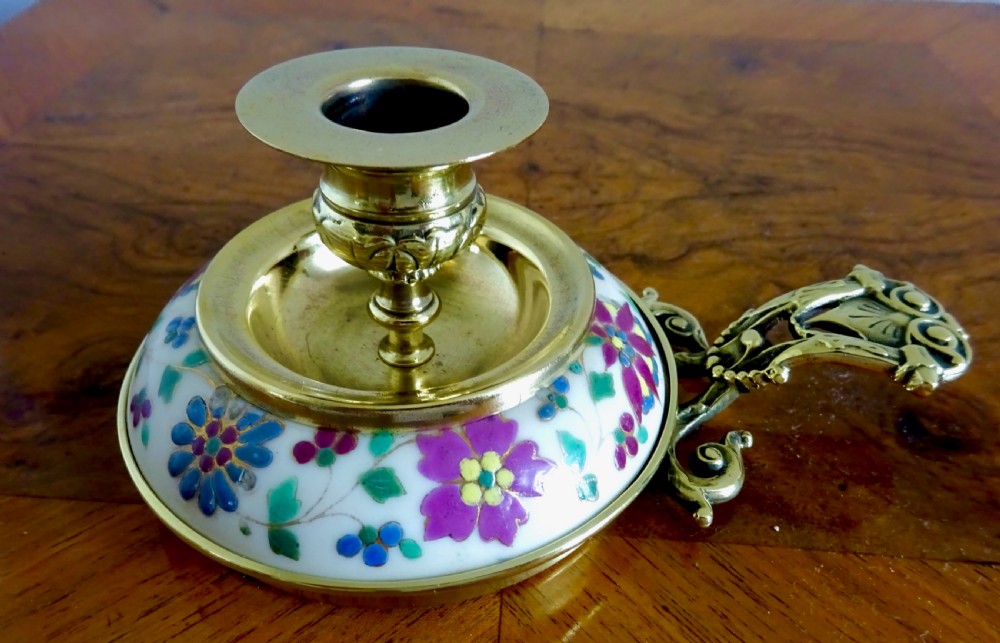 An Edwardian brass chamberstick with an ornate cast handle and a circular base decorated with floral enamel patterns. View VI