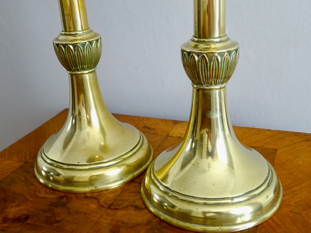 A pair of tall, polished brass candlesticks featuring turned shafts, carved leaf motif knops, circular bases, and wide upper sconces. View VII
