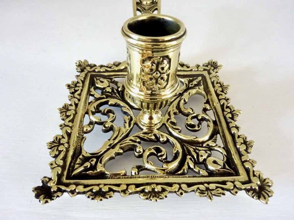 An antique brass chamberstick featuring a square openwork base with scrolling foliate patterns, a central cylindrical candle nozzle, and a long decorative handle with a mask detail. View IV