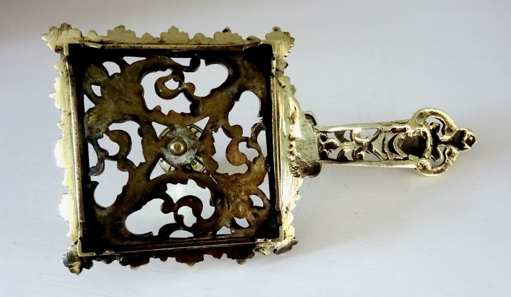 An antique brass chamberstick featuring a square openwork base with scrolling foliate patterns, a central cylindrical candle nozzle, and a long decorative handle with a mask detail. View V