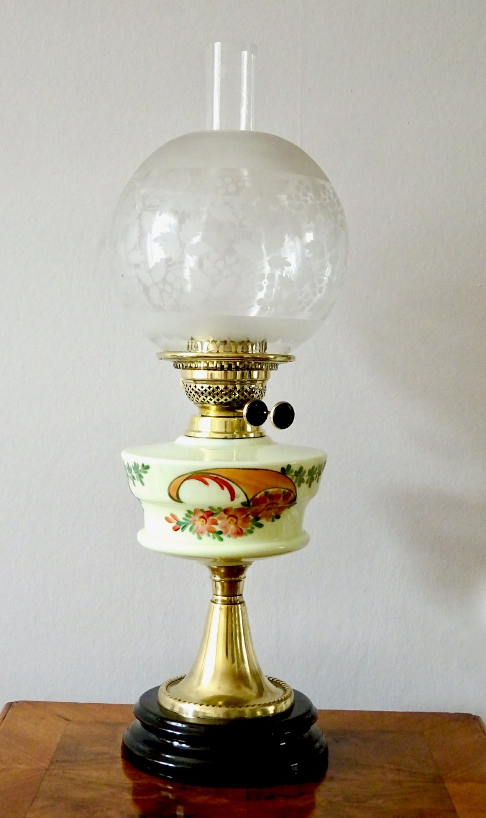 An antique brass oil lamp featuring a hand-painted floral glass font, a frosted etched globe shade, and a tall clear glass chimney on a stepped circular base.