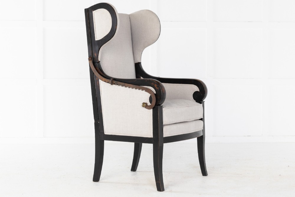 Early 19th-century French wingback armchair featuring an ebonised wooden frame, iron ratchet reclining mechanism, and light grey linen upholstery.