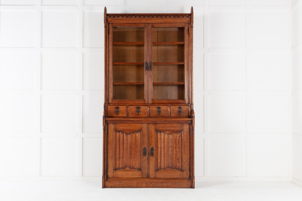 A tall nineteenth-century solid oak glazed bookcase featuring upper glass doors, four central drawers, and lower cupboard doors with carved linenfold panels.