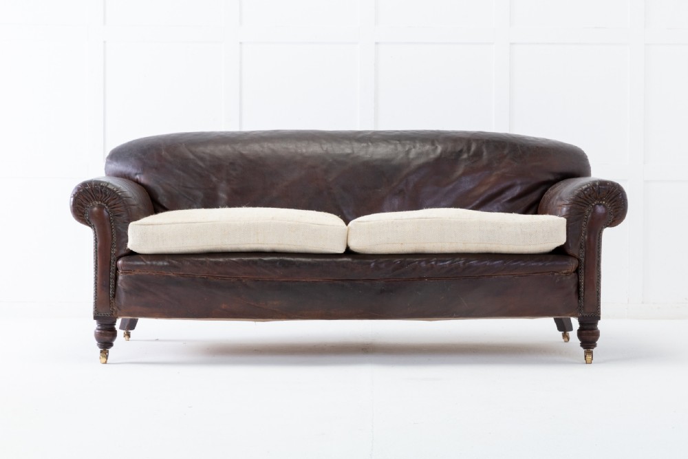 A two-seater 1920s English sofa featuring dark brown leather upholstery, rolled arms with studding details, light cream linen seat cushions, and turned wooden legs on brass castors.