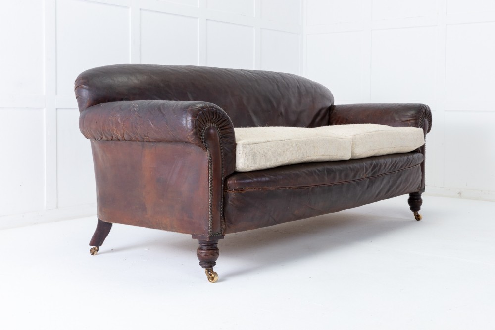 A two-seater 1920s English sofa featuring dark brown leather upholstery, rolled arms with studding details, light cream linen seat cushions, and turned wooden legs on brass castors. View II