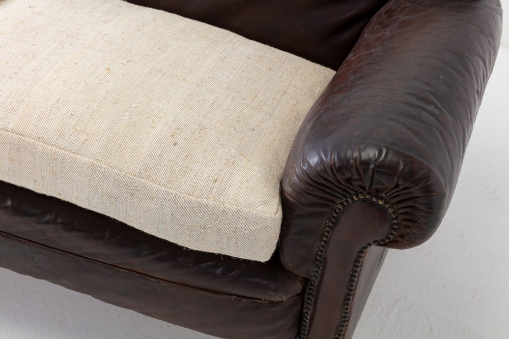 A two-seater 1920s English sofa featuring dark brown leather upholstery, rolled arms with studding details, light cream linen seat cushions, and turned wooden legs on brass castors. View III