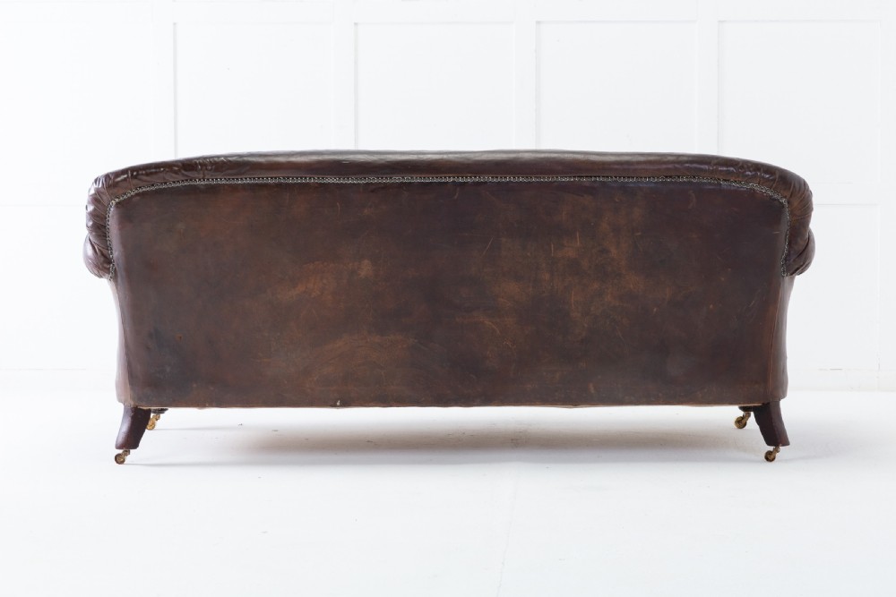 A two-seater 1920s English sofa featuring dark brown leather upholstery, rolled arms with studding details, light cream linen seat cushions, and turned wooden legs on brass castors. View IV