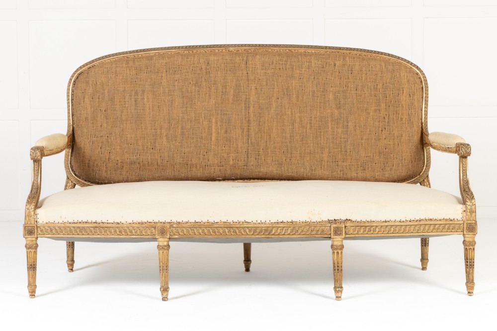 A 19th-century French Louis XVI style giltwood sofa featuring a carved gold leaf frame, fluted legs, a woven back panel, and light cream upholstered seating.