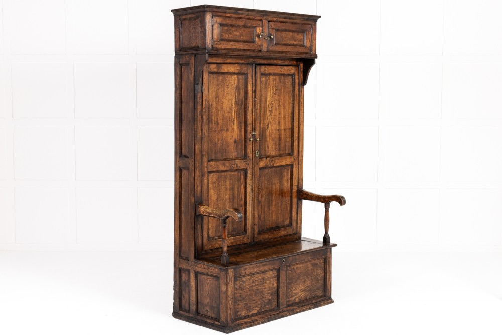 An 18th-century George III dark elm bacon settle featuring a tall panelled cupboard back, an upper hood cabinet, raised armrests, and a box seat base.