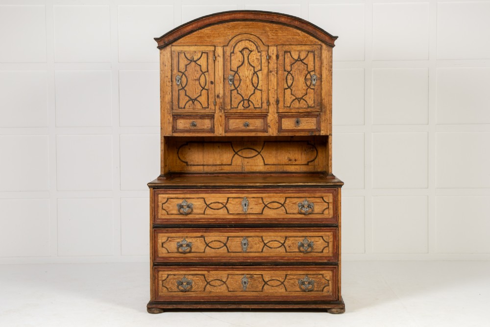 An 18th-century two-part pine cabinet featuring an arched upper cupboard section decorated with black painted geometric scrollwork over a base with three wide drawers.