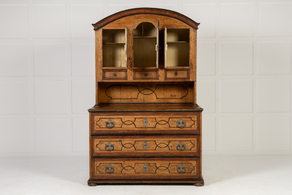 An 18th-century two-part pine cabinet featuring an arched upper cupboard section decorated with black painted geometric scrollwork over a base with three wide drawers. View II