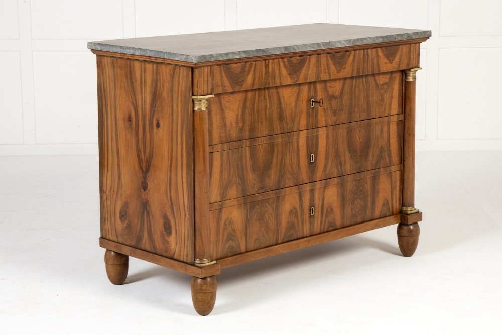 A 19th-century Italian walnut commode featuring a grey marble top, three drawer fronts with bookmatched grain, flanking column pilasters with brass mounts, and turned feet.